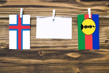 Hanging flags of Faroe Islands and New Caledonia attached to rope with clothes pins with copy space on white note paper on wooden background.Diplomatic relations between countries.
