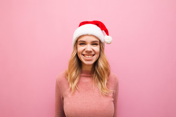 Portrait of smiling girl in christmas hat on pink background, looks into camera and laughs. Joyful lady santa is smiling, wearing a pink warm sweater and santa hat. Christmas concept