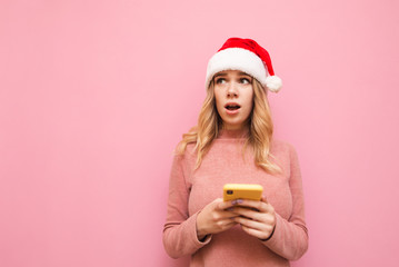 Portrait of shocked girl in pink sweater and Santa hat uses smartphone on pink background and looks surprised at blank space, wearing Christmas hat. Copyspace. X-mas concept
