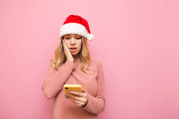 Portrait of surprised girl in pink sweater and Santa hat uses smartphone on pink background, looks into screen with surprised face. Emotional girl uses smartphone at Christmas. Isolated. Xmas concept