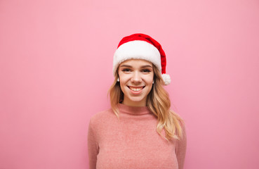 Portrait of a happy girl in a Santa Claus hat stands on a pink background, listens to music in wireless headphones and looks into the camera with a smile on his face. Girl listening to Christmas music