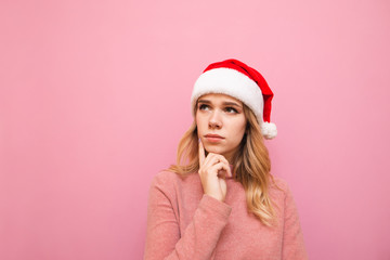 Closeup portrait of a cute girl in a Christmas hat isolated on a pink background, thoughtfully looking away with a serious face. Pensive lady in santa hat looks at empty space.