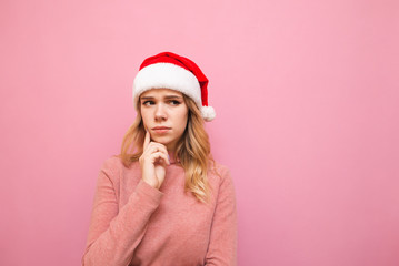 Attractive blonde in a hat santa looks thoughtfully aside at copy space, touches a finger to the cheek. Cute girl in a pink sweater and Christmas hat looks away thoughtfully. Christmas cocnept. xmas