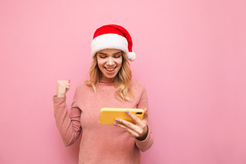Portrait of happy girl in christmas hat playing mobile games on smartphone, joyful rejoices to win on pink background, looks at smartphone screen and smiles. Christmas and New Year concept.