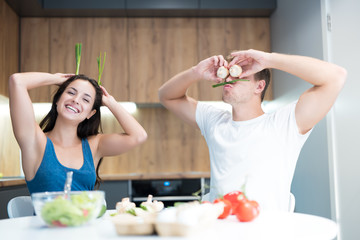 Obraz na płótnie Canvas young funny couple cooking breakfast in the kitchen man closes his eyes with mushrooms while his beloved wife holds green onion like ears laughing together