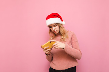 Concentrated gamer girl in christmas hat playing mobile games on smartphone on pink background. Portrait of focused teen girl in santa hat playing games on phone. Christmas gaming. X-mas