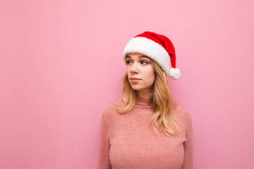 Beautiful girl in christmas hat and pink sweater looks away with serious face isolated on pink background. Christmas portrait of a cute girl in a santa hat. X-mas