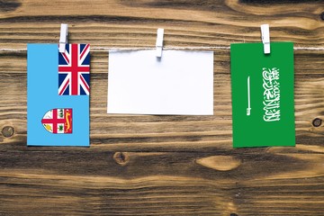 Hanging flags of Fiji and Saudi Arabia attached to rope with clothes pins with copy space on white note paper on wooden background.Diplomatic relations between countries.