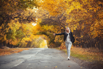 Obraz premium young woman stands and waits on an asphalt road in cold autumn weather, a girl tries to get from a suburban highway by a hitchhiker showing a hand gesture
