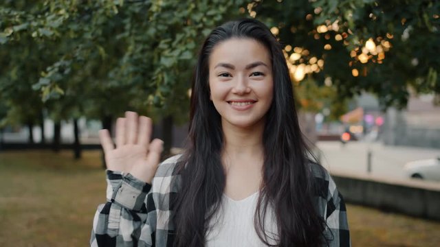 Slow motion of young Asian woman waving hand smiling looking at camera outside in city park. Millennials, positive emotions and happy youth concept.