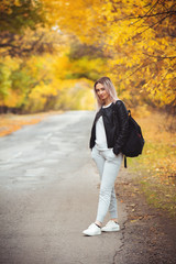 young woman in casual sportswear with a backpack standing on the asphalt road of a suburban road, girl travels walking and enjoying the autumn nature