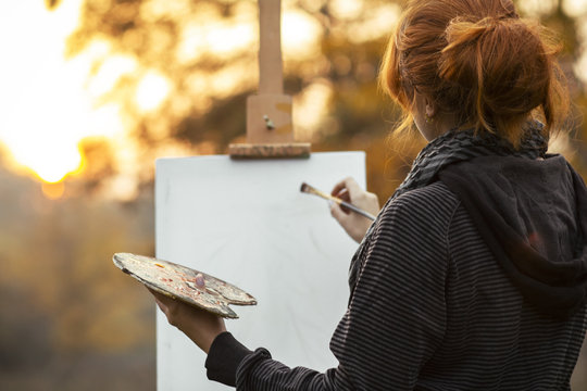back head of red-haired girl with a bun painting a picture on an easel in nature, a young woman engaged in art and enjoying beautiful landscape at sunset ,concept hobby and lifestyle