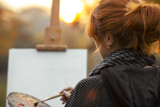 back head of red-haired girl with a bun painting a picture on an easel in nature, a young woman engaged in art and enjoying beautiful landscape at sunset ,concept hobby and lifestyle