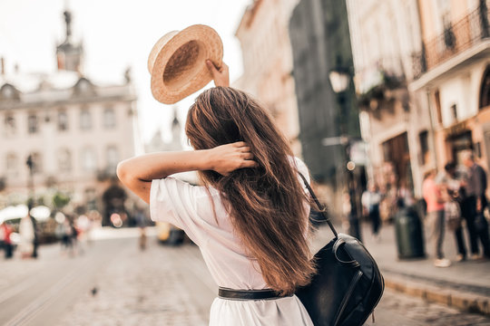 Young stylish woman walking on the old town street, travel with backpack, straw hat, wearing trendy outfit.