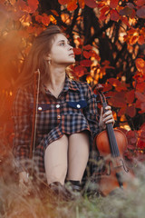 autumn portrait of beautiful young woman sitting on the ground with a violin on a background of red autumn leaves and looking up, concept of hobby and music