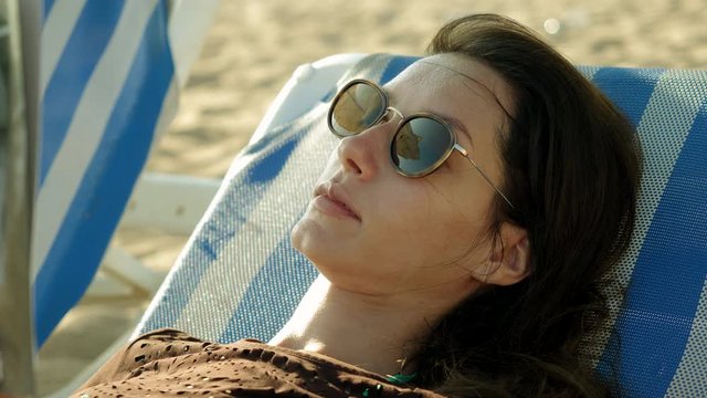 Coast of Costa del Maresme. Close-up shot of beautiful young woman spending the day on the beach, lying under a beach umbrella on a sun lounger and reading a book. Calella. Spain. 4K