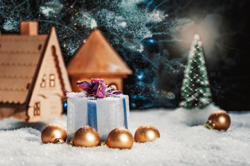 Christmas still life, gift box with a bow in the snow near the house and Christmas tree at night.