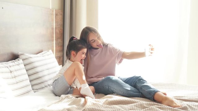 Two beautiful girls having fun in bedroom, attractive woman takes photo using cell phone, showing tongue full of joy, background of small pillows, morning time concept