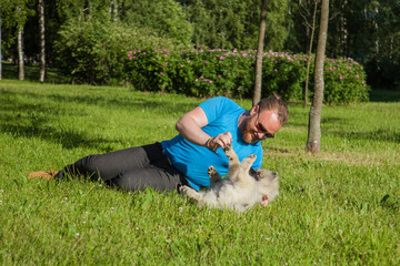 Young man with a red beard lay down on the grass in the park and plays with the keeshond / Wolfspitz puppy