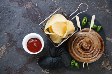 Stone slate with barbecued rolled sausages, potato chips and ketchup. Flatlay over brown stone background with copyspace