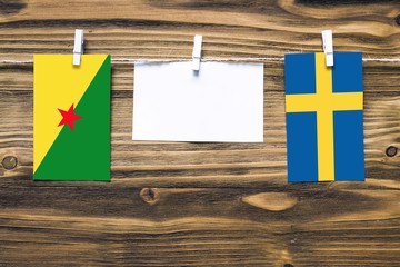 Hanging flags of French Guiana and Sweden attached to rope with clothes pins with copy space on white note paper on wooden background.Diplomatic relations between countries.