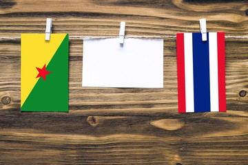 Hanging flags of French Guiana and Thailand attached to rope with clothes pins with copy space on white note paper on wooden background.Diplomatic relations between countries.