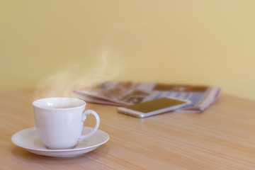 White coffee cup and soft stream smoke put on wood table with blurry smartphone and newspaper. Business man have a breakfast in a cozy room and dark yellow cement wall with radiant sunrise background.