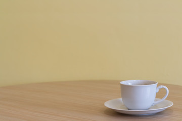 Obraz premium White coffee or tea cup with not soft stream smoke put on wooden table in a living room and blurry dark yellow cement wall with window lighting background. Selective focus and copy space.