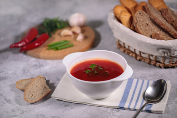 Tasty and hearty dinner. A plate with borsch on the table, next to the board is parsley, dill, green onions, garlic, chili pepper and a basket with bread.