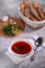 A plate with beetroot borsch on the table, next to the board is parsley, dill, green onions and garlic and a basket with bread. Tasty and hearty lunch.