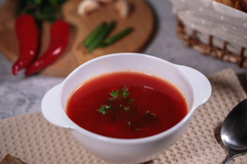 Tasty and hearty dinner. A plate with borsch on the table, next to the board is parsley, dill, green onions, garlic, chili pepper and a basket with bread.