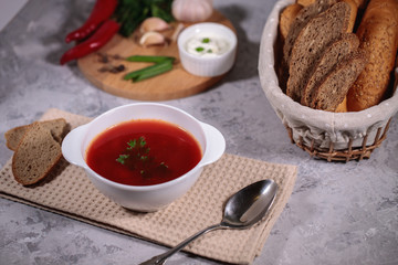 Tasty and hearty dinner. A plate with borsch on the table, next to the board is parsley, dill, green onions, garlic, chili pepper and a basket with bread.