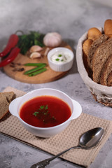 Tasty and hearty dinner. A plate with borsch on the table, next to the board is parsley, dill, green onions, garlic, chili pepper and a basket with bread.