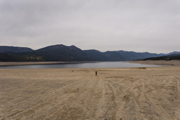Fototapeta na wymiar Single person standing on an empty beach of a drained lake with a mountain range in the background