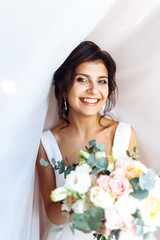 Young bride in white dress with a bouquet of flowers posing under curtain. Beautiful woman with professional make up and hair style. Morning of the bride. Wedding day. Fashion bride.