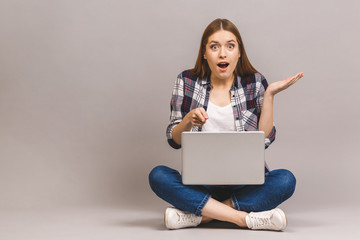 Wow, great news! Happy young amazed woman sitting on the floor with crossed legs and using laptop isolated on gray background.