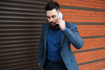 Stylish handsome brunette man with beard, wearing classic formal blue suit jacket, talking on a smartphone outdoors on the city street near garage roller door. Businessman using mobile phone.