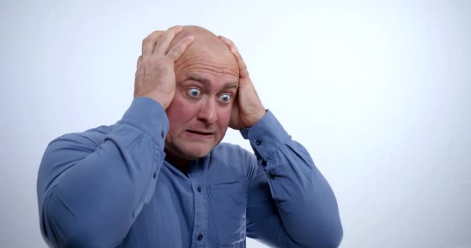 Close-up portrait of a nervous bald middle-aged man in a blue shirt, he is in the Studio on a white background, he rubs his head and face with his hands, terribly staring like a madman.