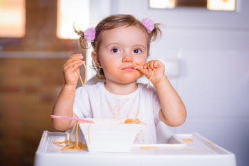 Cute little 1 year old girl, baby, child, sitting in a high-powered chair at home, eats noodles hands, photo in real life interior, self-feeding concept