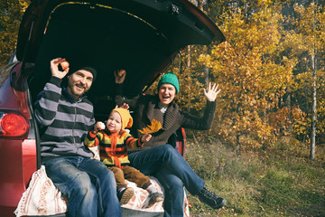 Happy family resting after day spending outdoor in autumn park. Father, mother and child sitting inside car trunk, smiling and looking to the camera. Travel in fall season.