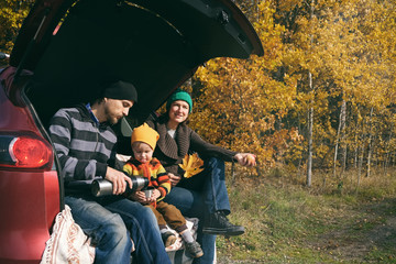Happy family resting after day spending outdoor in autumn forest. Father, mother and child sitting inside car trunk and drink tea from thermos. Travel in fall season.