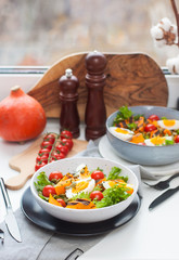 Lunch bowl with cherry tomatoes, roasted pumpkin, green salad, seeds and boiled eggs. Grey plate on white background. 