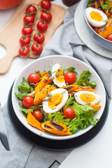 Lunch bowl with cherry tomatoes, roasted pumpkin, green salad, seeds and boiled eggs. Grey plate on white background. 