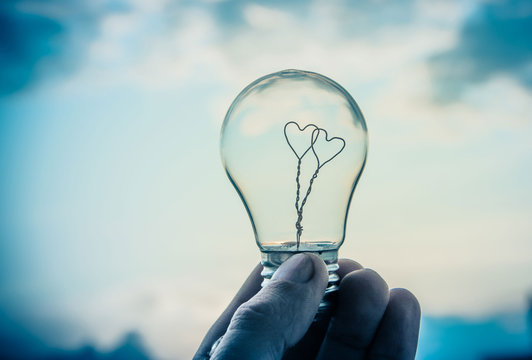 A man holds a light bulb in his hand, inside which there are two hearts against the sky and the sun. The energy of the Sun and wind, the concept of pure energy, love and unity with nature.