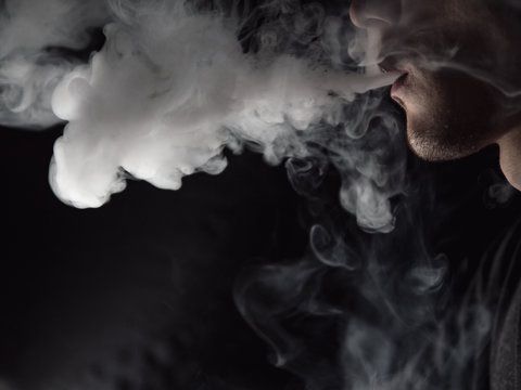 Close up of a male vaper exhaling a cloud of steam on a dark background