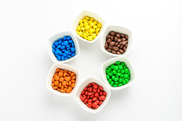 Six squared white bowls with small mixed colored chocolate coated candies, isolated on white background, top view or flat lay photo of yellow, orange, green, blue, red and brown sweets