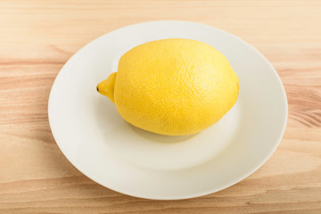 One fresh yellow lemon on a white plate on a wooden brown table, top view or flat lay photo of healthy fruit