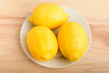 Three fresh yellow lemons on a white plate on a wooden brown table, top view or flat lay photo of healthy fruits