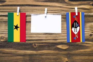 Hanging flags of Ghana and Swaziland attached to rope with clothes pins with copy space on white note paper on wooden background.Diplomatic relations between countries.