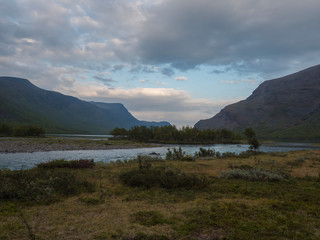 Beautiful wild Lapland nature landscape with blue glacial river, Kaitumjaure lake, birch tree forest and mountains. Northern Sweden summer at Kungsleden hiking trail. Blue sky dramatic sunset clouds.